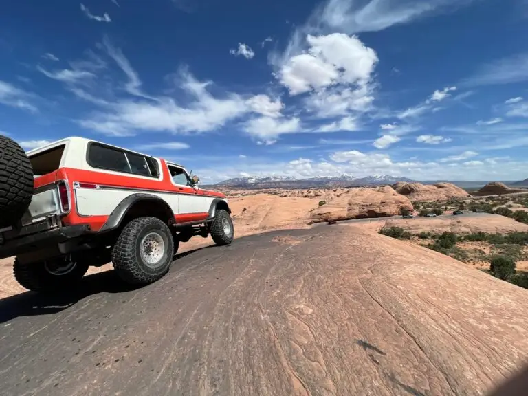 White and red early Bronco driving up a slickrock road under a bright blue sky