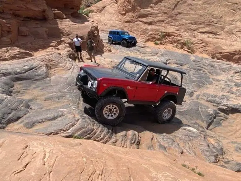Red early Bronco climbing a slickrock section with people watching