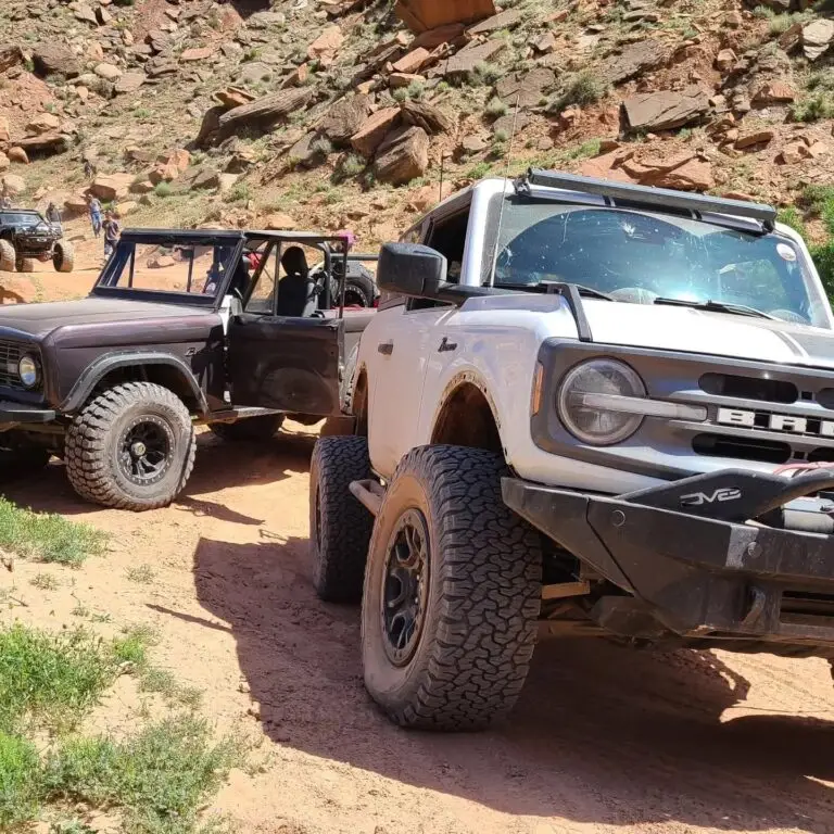 Black early Bronco and a silver and white newer Bronco parked on a red rock trail